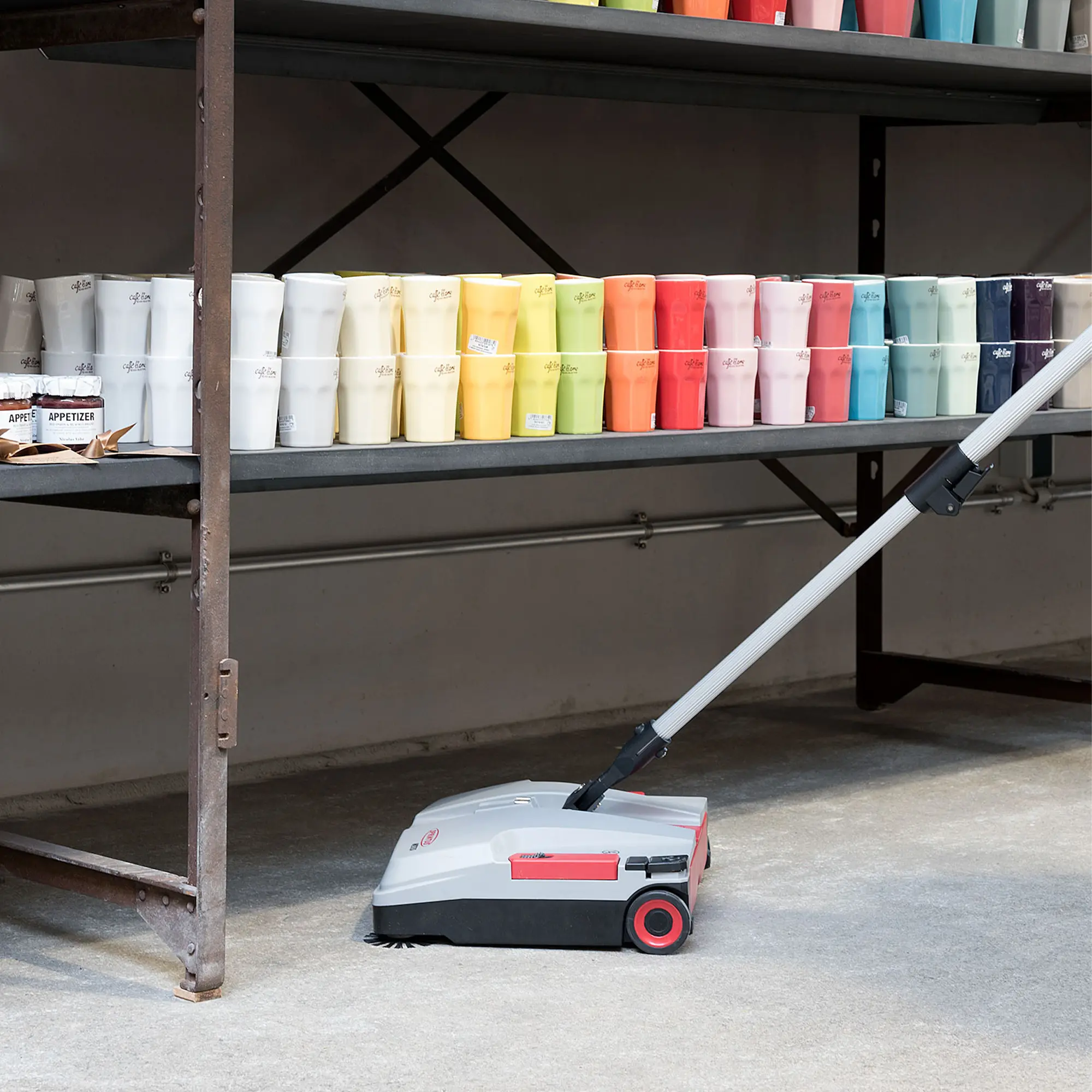 Sprintus Akkukehrmaschine Medusa zur kabellosen Zwischenreinigung von Hartboden in Lager oder Verkaufsraum mit Regalen und farbigen Bechern